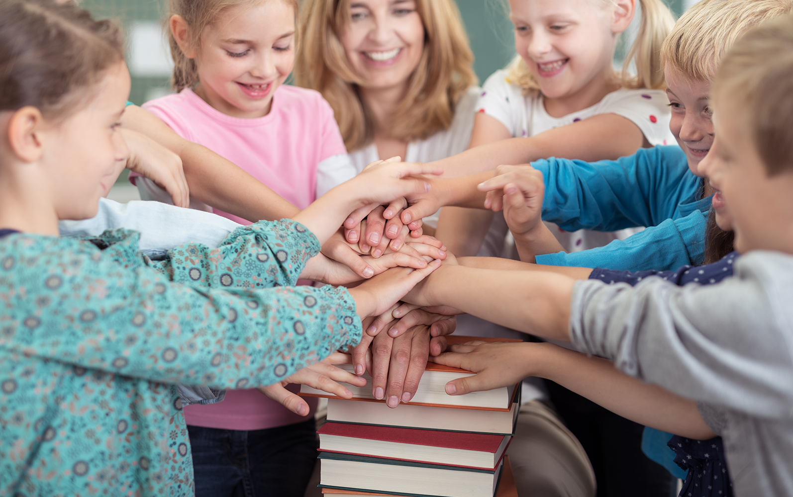 Mehrere Kinder und eine Lehrerin legen ihre Hände übereinander auf einen Bücherstapel