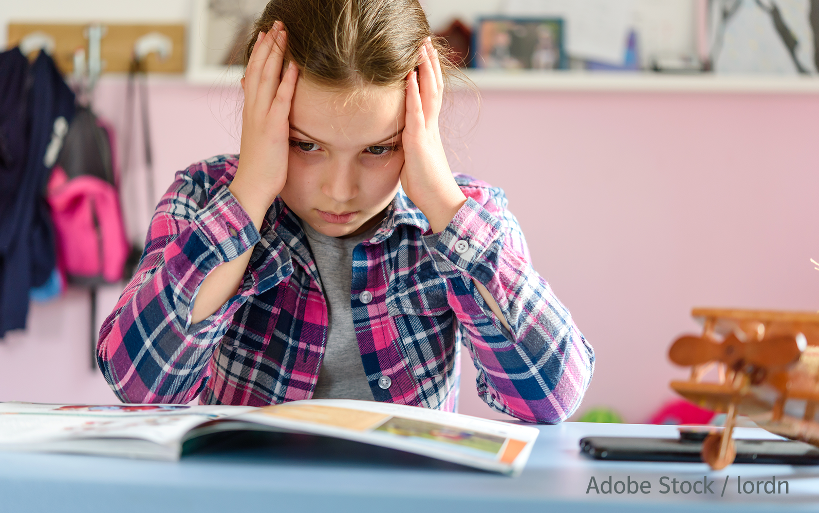 Ein Mädchen sitzt an einem Tisch und stützt den Kopf mit beiden Händen ab. Ihr Blick ist gesenkt auf ein Buch gerichtet