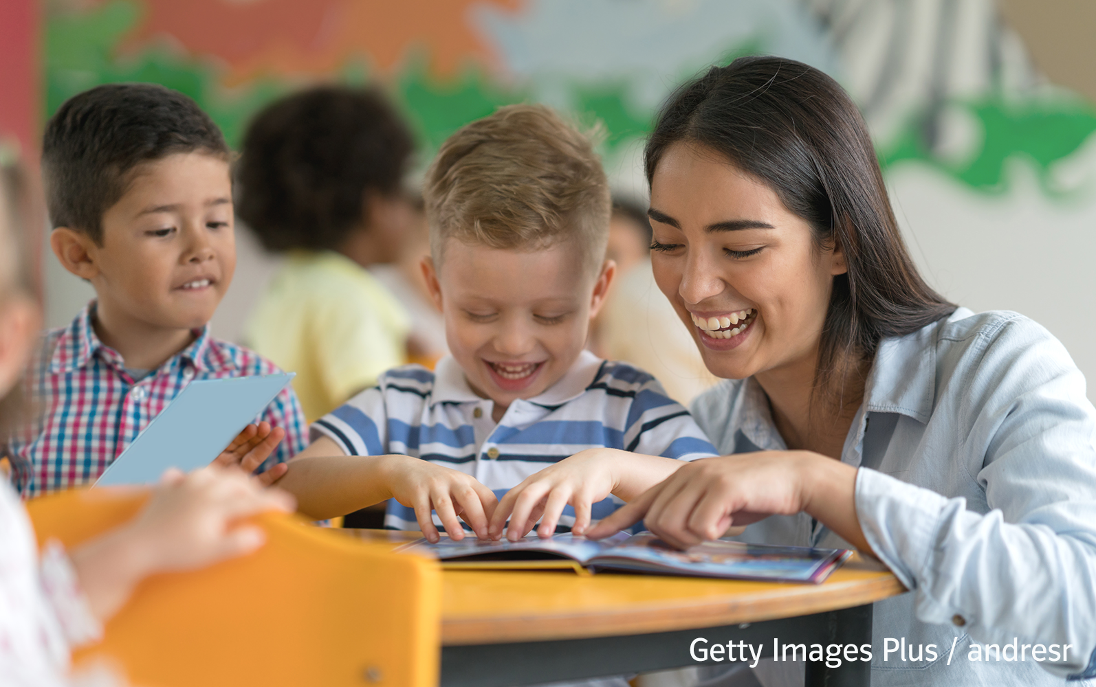Eine Lehrerin und zwei junge Schüler schauen in einem Klassenzimmer zusammen in ein Buch und lachen