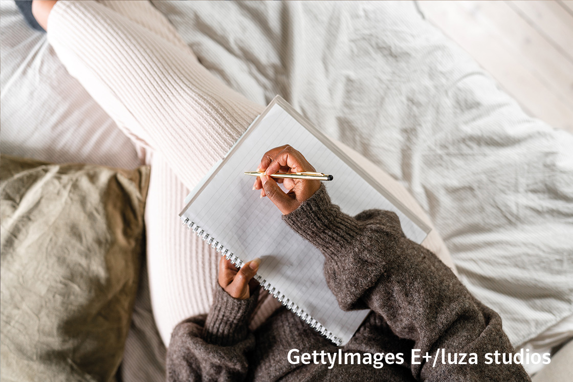 Eine Person schreibt in ein Notizbuch und sitzt auf einem Bett  