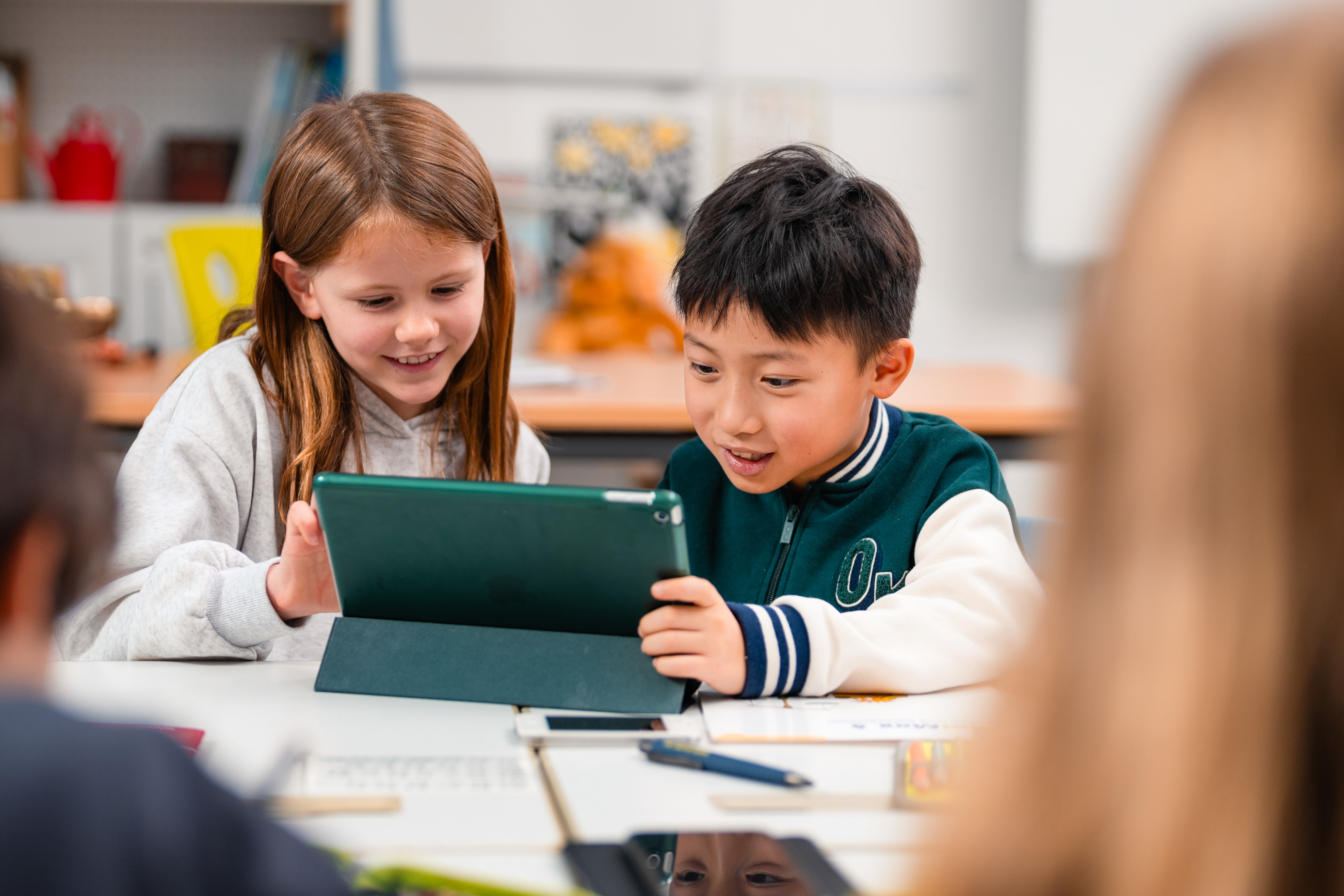 Zwei Lernende sitzen an einem Tisch in einem Klassenzimmer und schauen gemeinsam auf ein Tablet Zwei Lernende sitzen an einem Tisch in einem Klassenzimmer und schauen gemeinsam auf ein Tablet