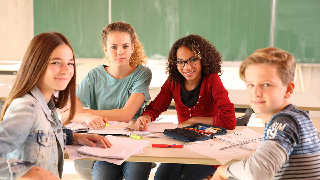 Vier Lernende sitzen an einem Tisch mit Stiften und Blöcken in einem Klassenzimmer und schauen in die Kamera
