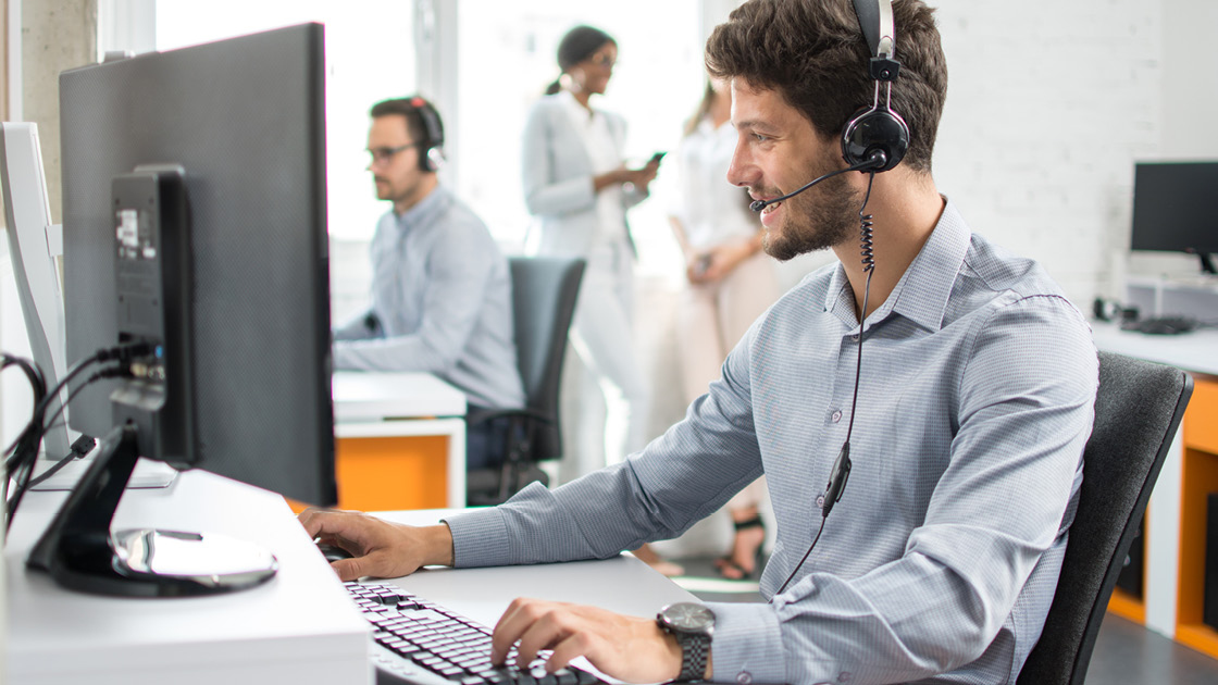 Ein Mann sitzt mit einem Headset an einem Computer an einem Schreibtisch in einem Büro