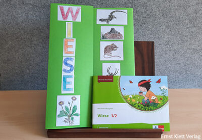 Gebasteltes Lapbook mit der Aufschrift „Wiese“ und davor das Cover des Anoki-Übungshefts „Wiese 1 Schrägstrich 2“