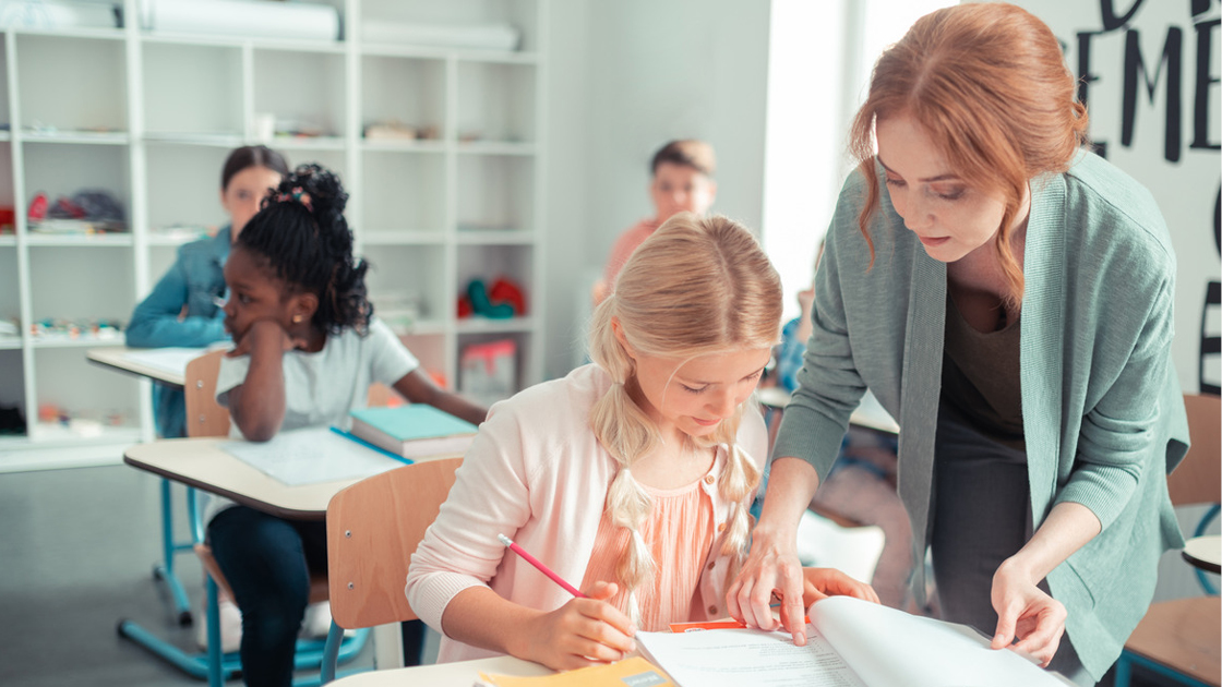 Eine Lehrkraft hilft einer Schülerin an einem Schreibtisch. Weitere Schüler sitzen an Tischen