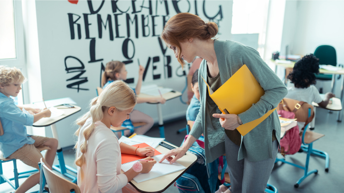 Eine Lehrkraft hilft einer Schülerin an einem Schreibtisch. Weitere Schüler sind mit Aktivitäten beschäftigt 