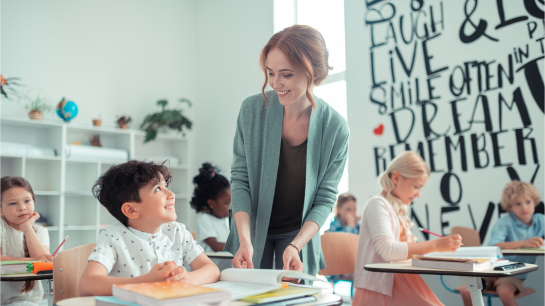 Eine Lehrkraft hilft einem Schüler an einem Schreibtisch. Weitere Schüler sind mit Aktivitäten beschäftigt 