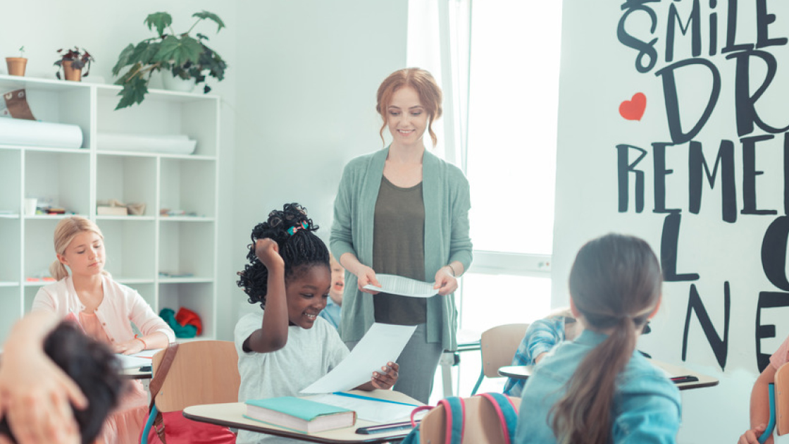 Eine Lehrerin steht neben einer Schülerin, die ihre Hand erfreut hebt. Weitere Lernende sitzen an Tischen