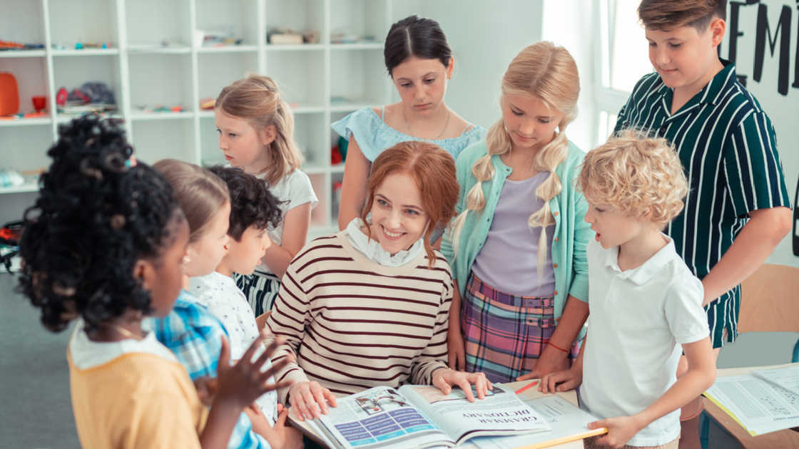 Eine Gruppe von Kindern und eine Lehrerin sitzen um einen Tisch mit einem Buch in einem Klassenzimmer