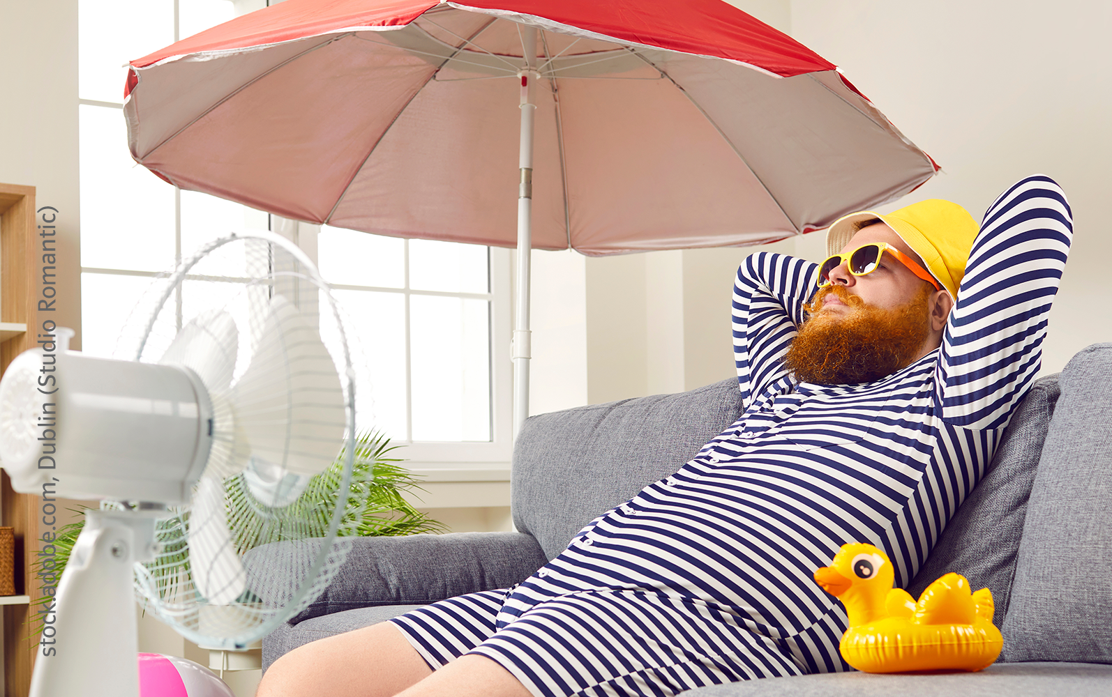 Mann im gestreiften Schwimmanzug entspannt auf Sofa mit Sonnenschirm und Ventilator