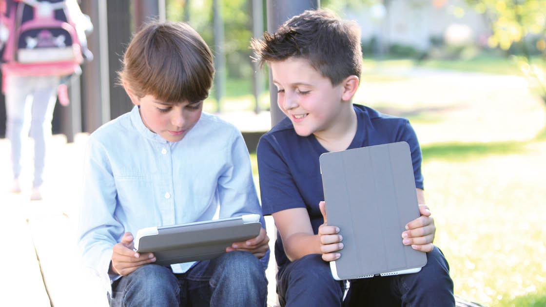 Ein Junge sitzt auf dem Schulhof und bedient ein Tablet, ein anderer sieht lächelnd zu