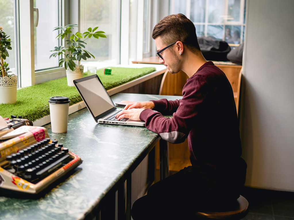Student sitzt mit Laptop und einem Pappbecher Kaffee in einem Café