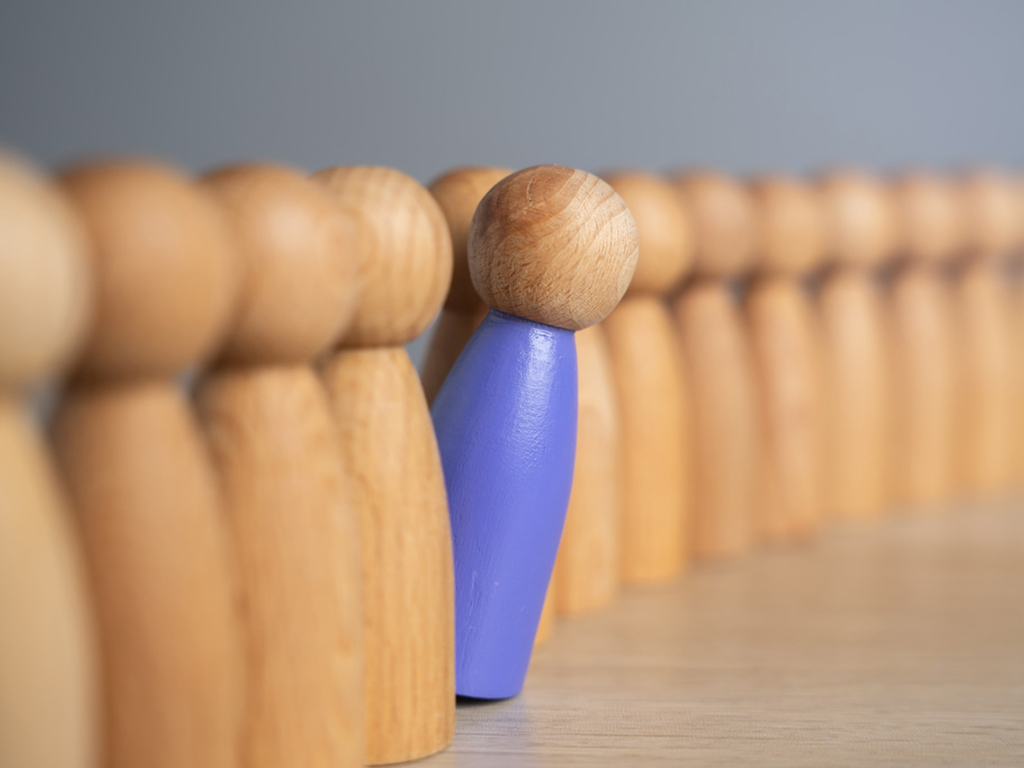Kleine braune Holzfiguren stehen hintereinander, eine lila Holzfigur verlässt die Reihe