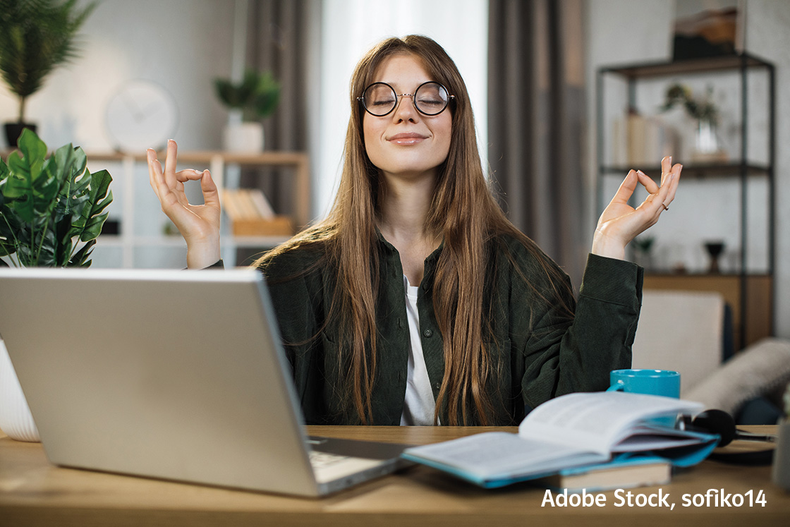 Eine Frau sitzt an einem Schreibtisch mit einem Laptop und meditiert mit ihren Händen in einer Mudra-Position