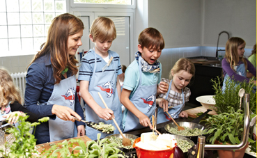 Eine Frau und mehrere Kinder kochen gemeinsam in einer Küche. Sie sind um einen Herd versammelt und bereiten Essen zu